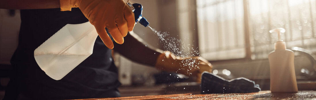 A gloved hand spraying disinfectant onto a wooden tabletop surface